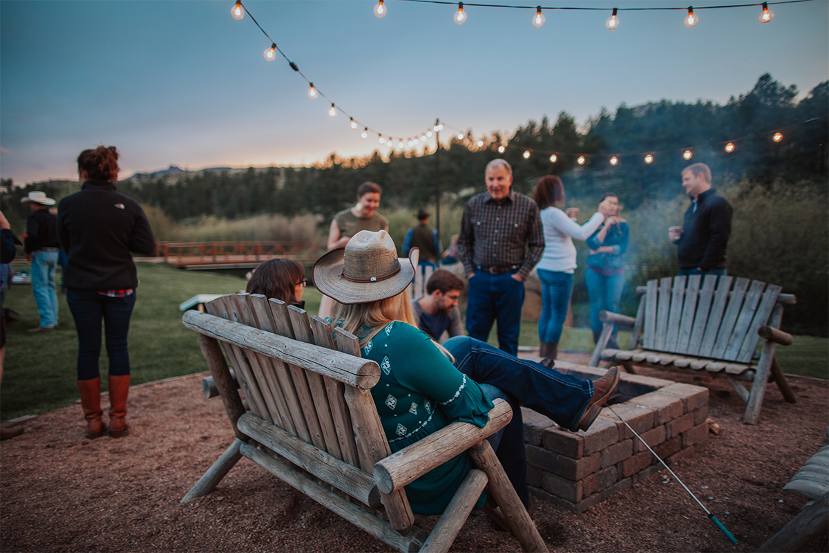 Adults mingle around a brick firepit at Lost Valley Ranch. The sky is darkening and overhead string lights begin to glow.