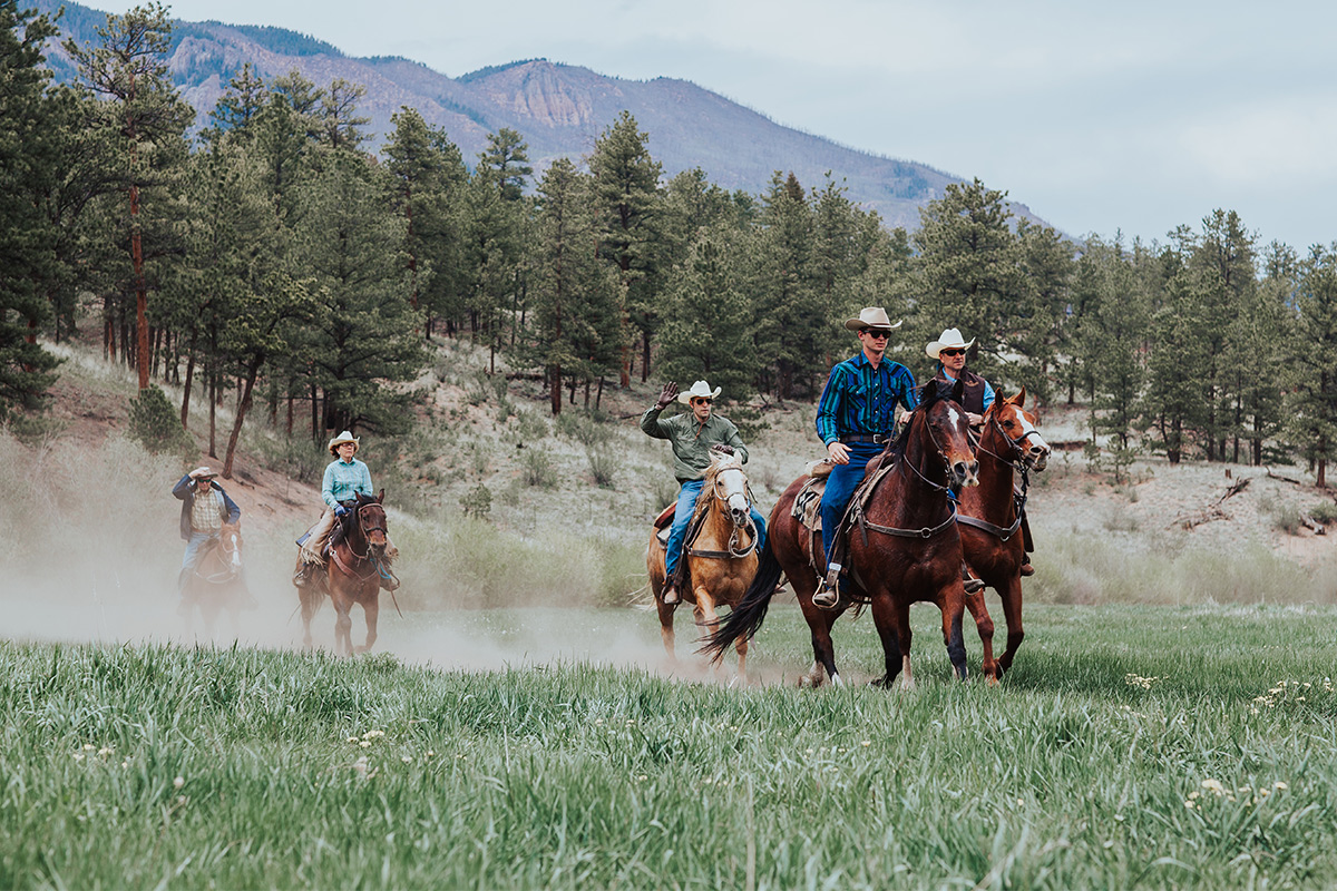 A group of people ride horses through a field near Lost Valley Ranch. Behind them is a lush pine tree forest and mountain peaks.
