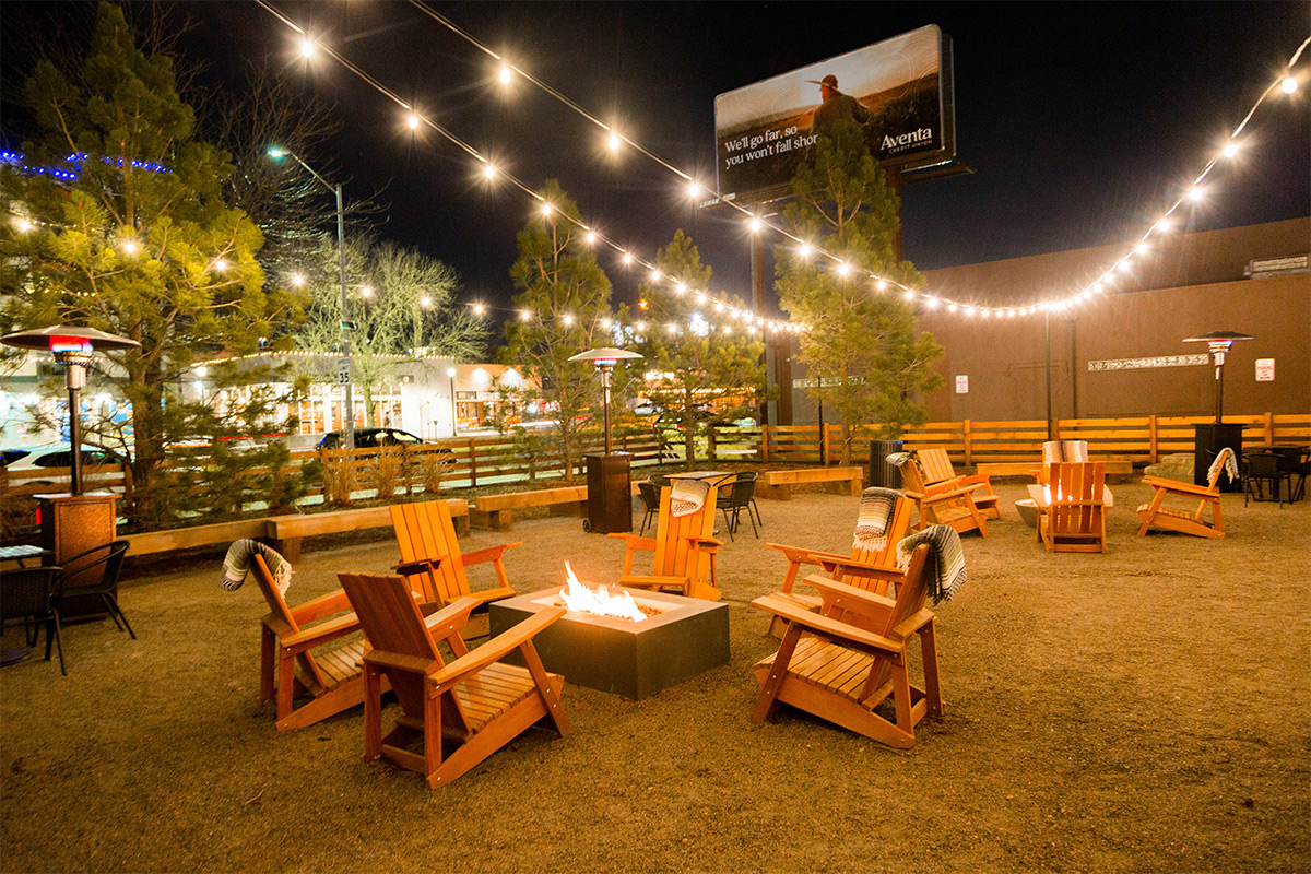 String lights hang over a roaring firepit with four wooden Adirondack chairs outside at The Yard.