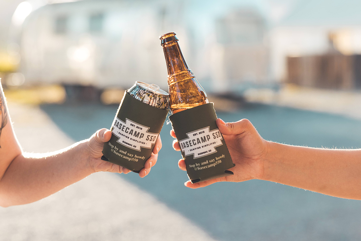 Two people clink alcoholic beverages in Basecamp 550 koozies outside near a metal camper.
