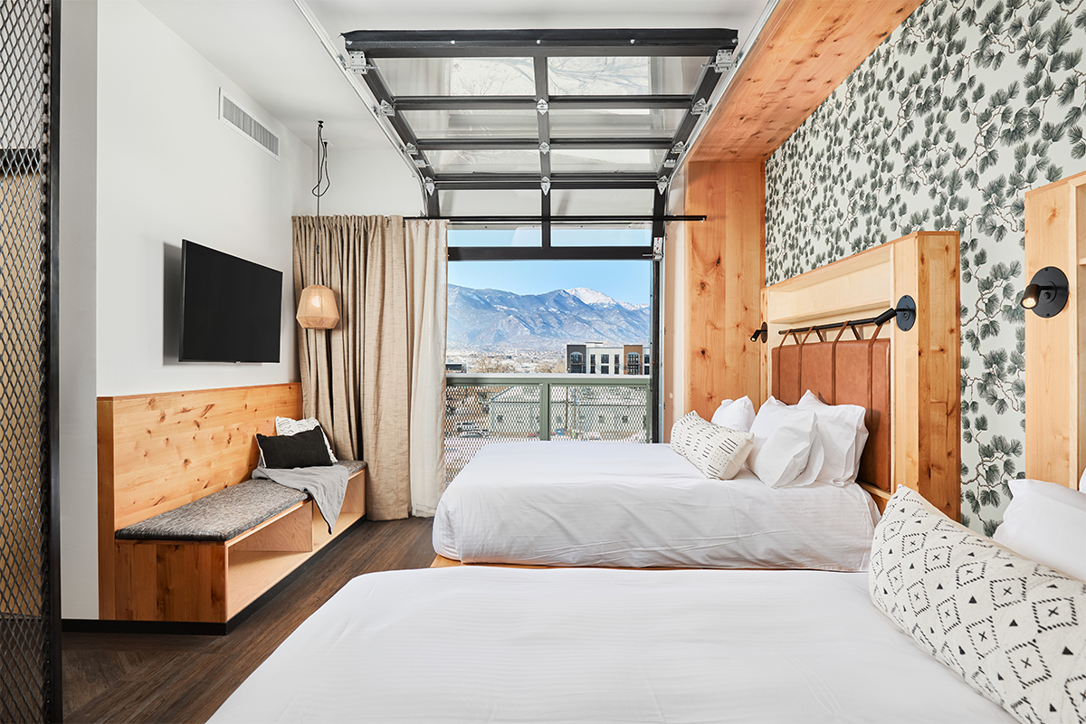 Two neat queen beds await guests in a suite at Kinship Landing. The garage-style window is open and mountain are visible in the distance.