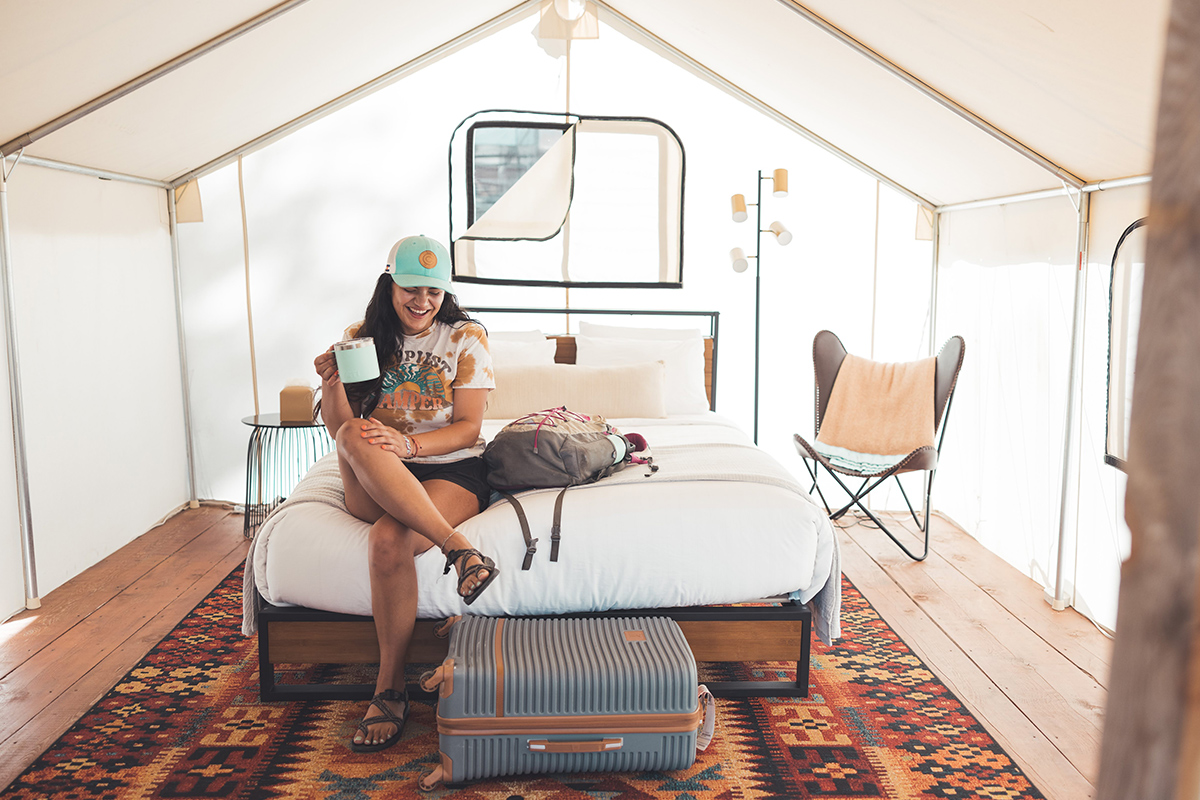 A guest wearing a robin-egg blue hat and holding a matching portable coffee mug sits on the bed with their luggage in a glamping tent at Basecamp 550.