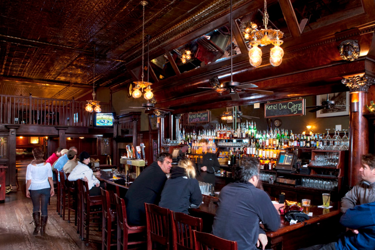 Guests sit and order drinks at a dark-wood bar with tavern-style exposed-bulb lighting overhead at the New Sheridan Hotel in Telluride, Colorado.
