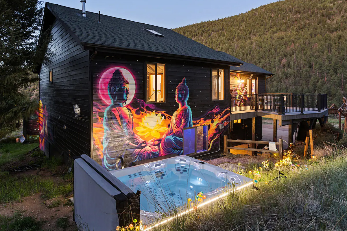 Small lights glow alongside a large uncovered hot tub at Hummingbird Hill near Idaho Springs, Colorado. Nearby a Buddhist-style mural is painted on the side of a black wall.