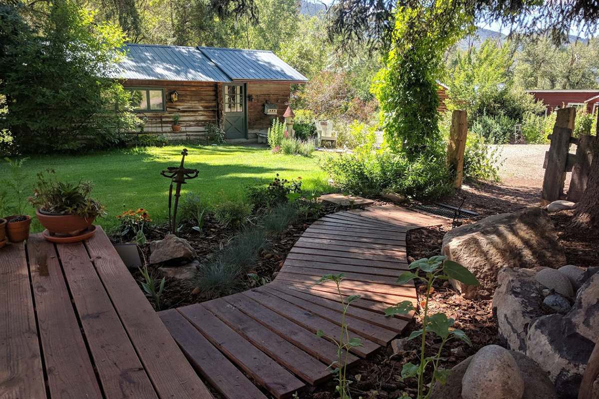 Smooth wooden planks create a path through a verdant garden to a raised porch Four Mile Creek B&B. In the background is a guest log cabin.