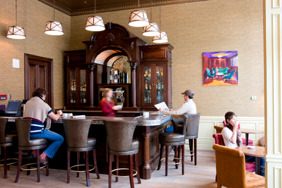At the parlor of the New Sheridan Hotel, two people sit at bar in high-back leather stool while a wait staff member in a red shirt brings an order.