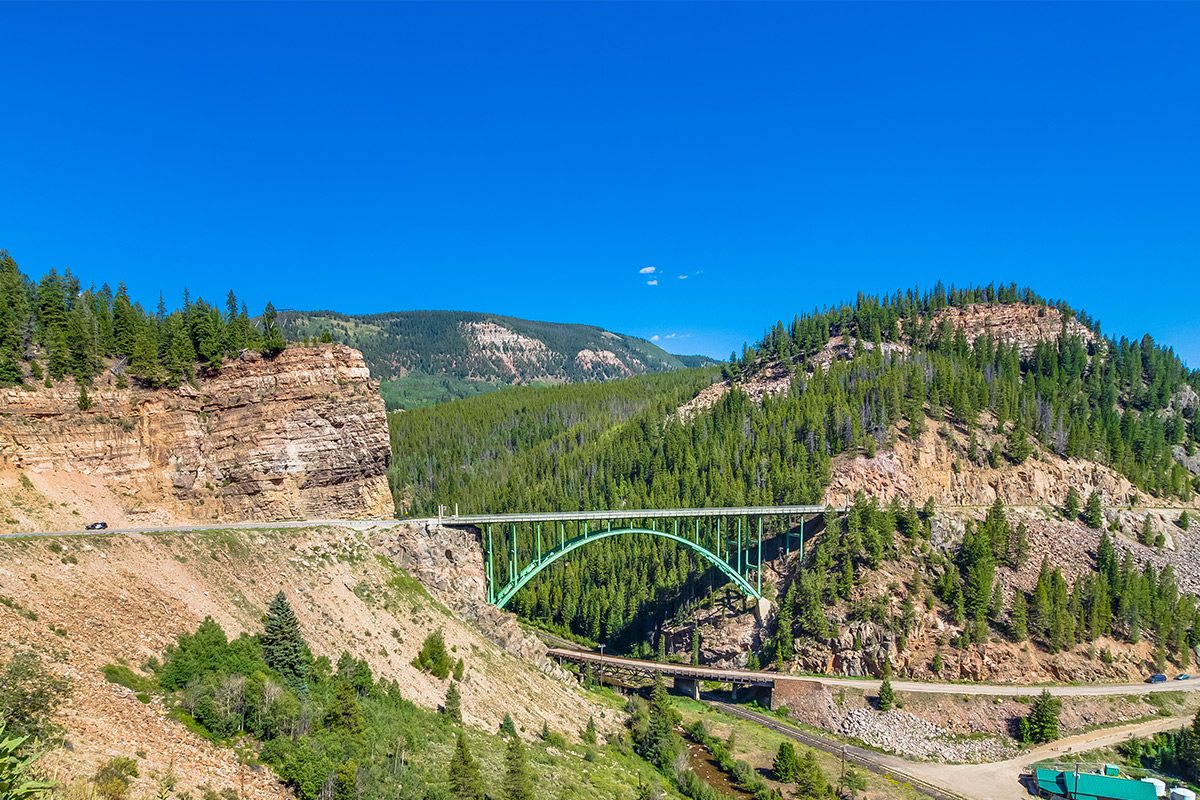 A strong, green bridge connects to sides of a river valley near Red Cliff, Colorado. Both sides of the valley are rocky and forested.