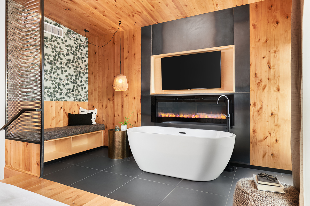 A modern, white bathtub is set in front of a gas fireplace. The room around the tub is decorated in black tile, wood paneling and wallpaper.