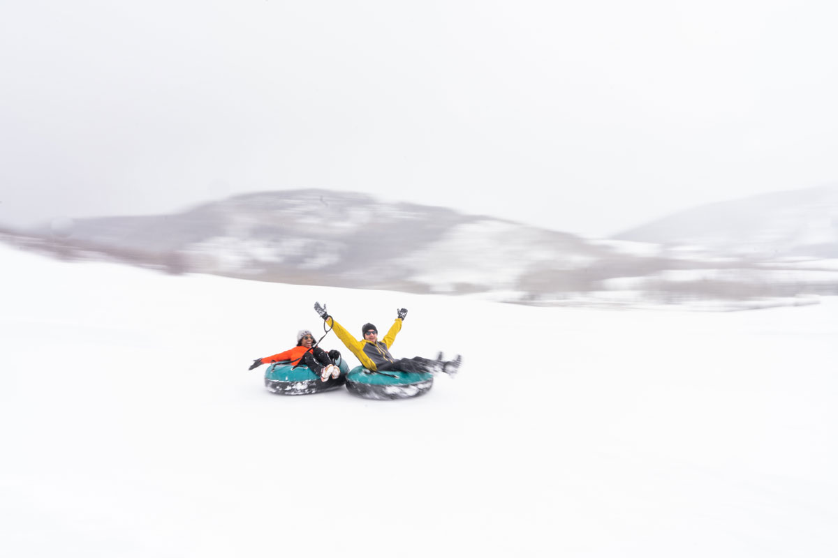 Two laughing and smiling people in their respective blue-and-black snow tubes tied to each other slide down a white, snowy hill as it snows outside. One of the people throws their hands up in the air looking at the camera, and the person behind them has their arms out wide beside them, looking at the person in front.