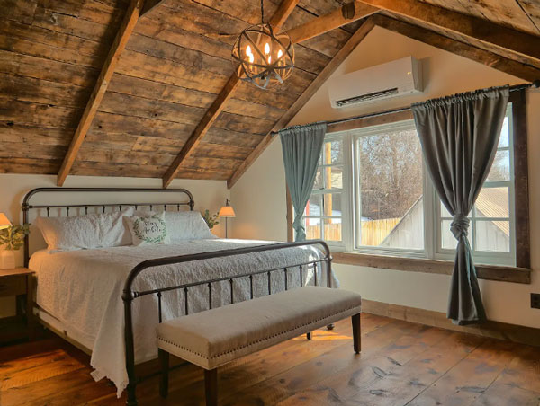 A cozy, wood-ceilinged and -floored bedroom at the Dyer Inn in Castle Rock. From the ceiling, a circular chandelier hangs above the large, clean, metal-framed bed. There is a large three-paned window overlooking some trees and draped with dusty blue thick curtains.