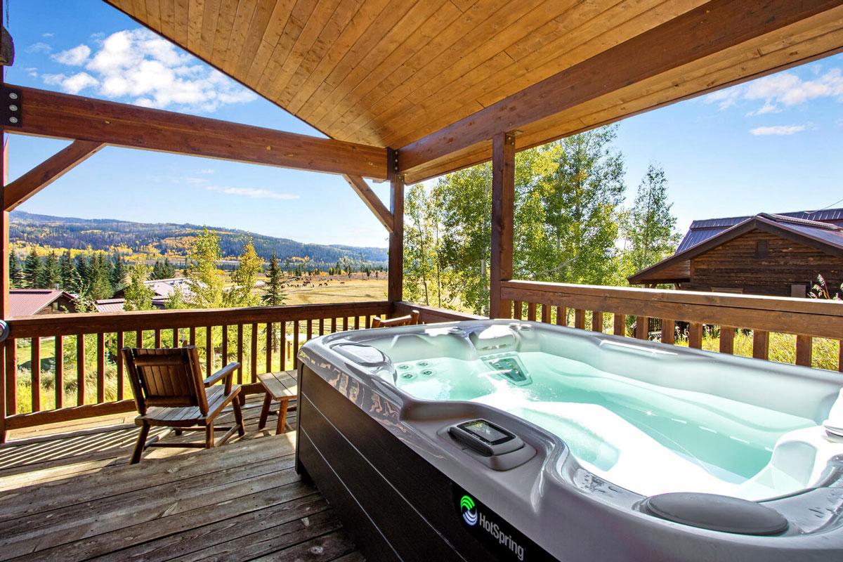 A large HotSpring hot tub filled with clear water sits on a deck in the sun at the Vista Verde Guest Ranch in Clark. The deck looks out on another adjacent cabin as well as the distant tree-spotted hills under a blue sky.