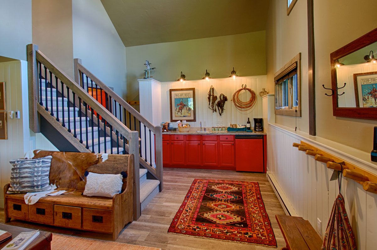 A warm-colored, large room with a small kitchen in the back of the photo. The cabinets in the kitchen are red and above the sink hangs a framed photo, horse-riding equipment and a rope for decoration. There are stairs leading up to another level. In front of the stairs is a wooden bench decorated with two throw pillows. The room is lit with natural light as well as light from the fixtures in the kitchen.