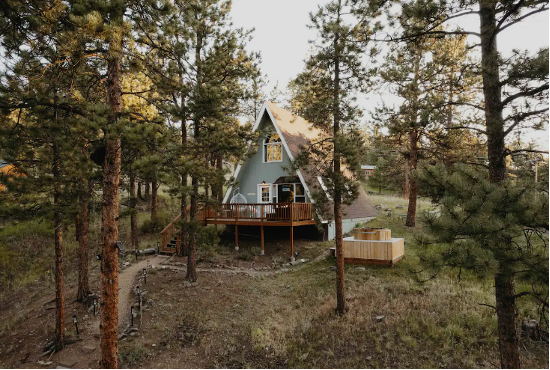 A mint-green Aframe in Bailey, Colorado, stands among pine trees with an open clearing in front of it.
