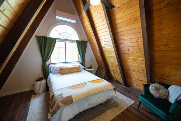 An image of the main bedroom in the Bailey A-Frame. The A-frame ceiling is dark brown wood, framing a large arched window with green curtains above the white-linen bed. There are little tables on either side of the bed. In the lower right corner of the photo is a dark green arm chair with a round pillow and a blanket thrown on the back.