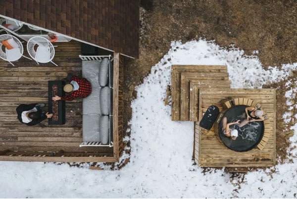 An aerial shot of the cedar hot tub and half of the patio at the Bailey A-Frame. There are two people in the hot tub and two people tending to the patio firepit on the patio. The patio is decorated with a few white wire chairs and a large gray couch. The brown ground is covered mostly in snow.