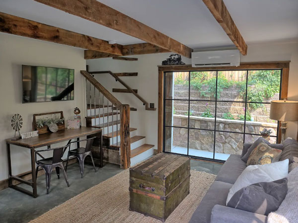 A spacious and cozy room with a large floor-to-ceiling window overlooking a green-leafed garden and stone garden walls. The room is decorated with a wooden desk, wooden accents on the ceiling, staircase and walls.