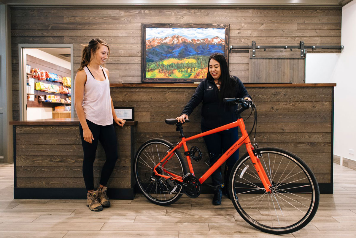Two people stand in the hotel's lobby. One person holds a big red bicycle upright while the other person in a tank top, leggings and hiking boots looks at it.
