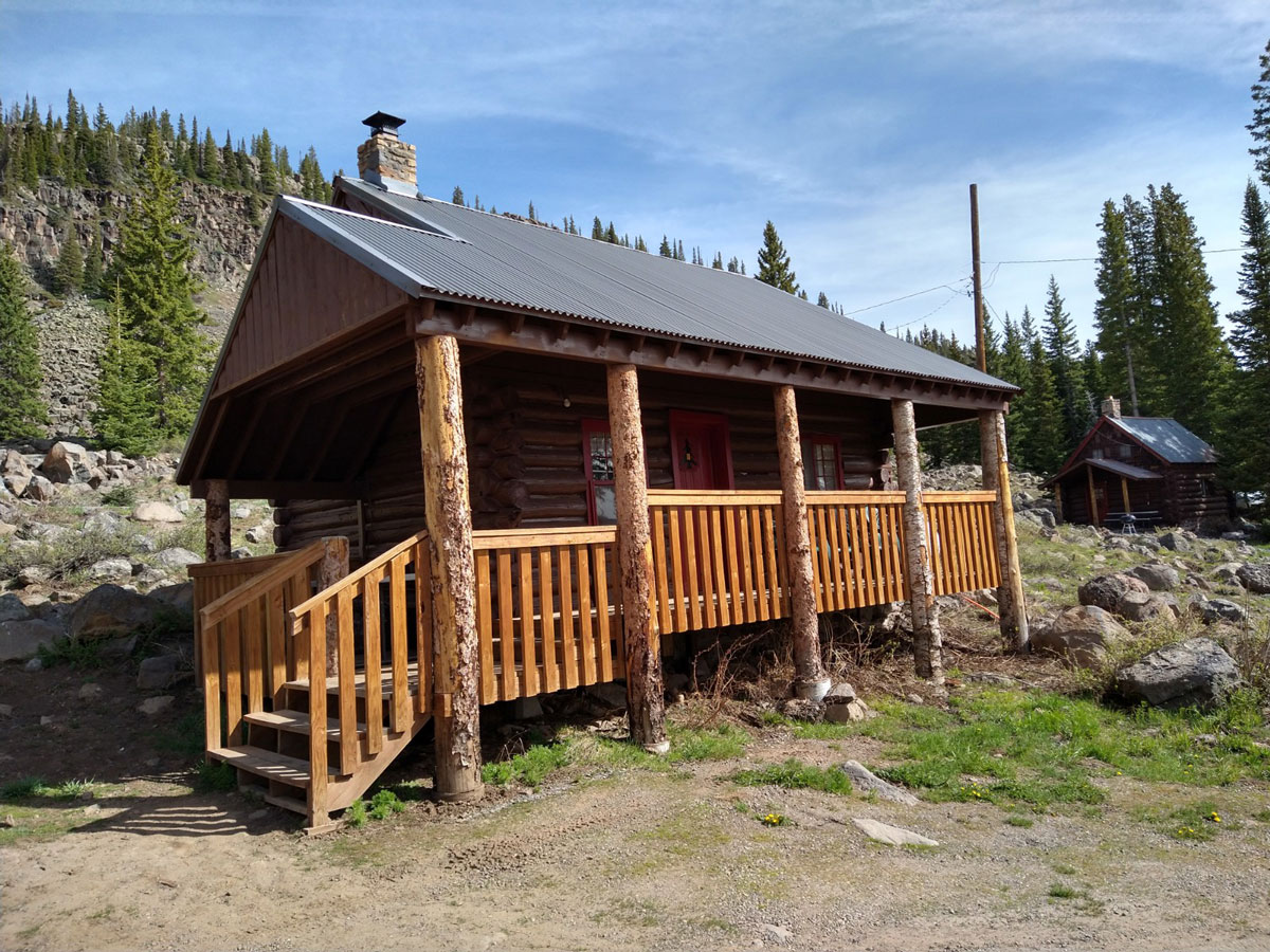 A rustic cabin is seen in the foreground with another in the background at Grand Mesa Lodge. The landscape is rocky, covered in brush, grass and evergreen trees.