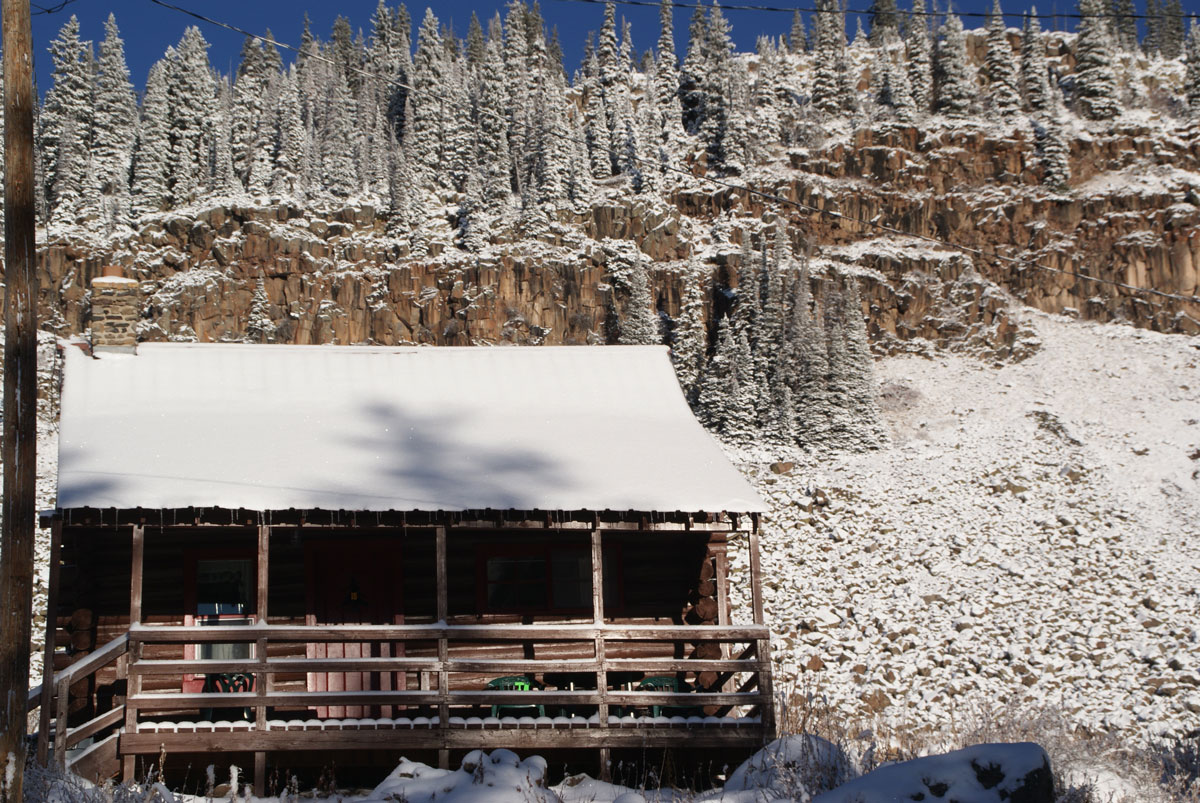 A rustic cabin at Grand Mesa Lodge is dusted with snow. The rugged landscape behind it is snowy, too, with a bright blue sky behind it.