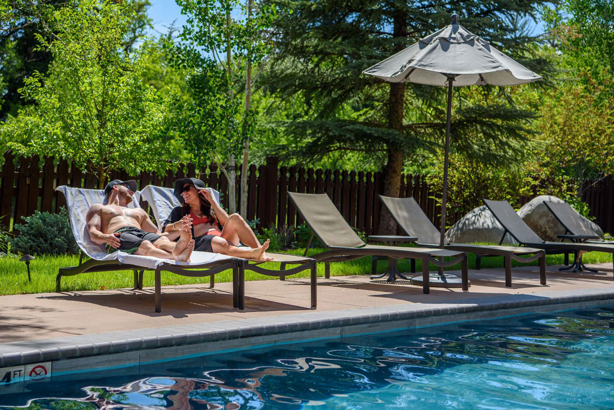 Two swimsuit-clad people hold hands and laugh together on pool chairs. There are tons of aspen and pine trees behind them and behind the short wooden fence outside of the pool. The sun is shining and reflecting off the small waves of the blue pool water.