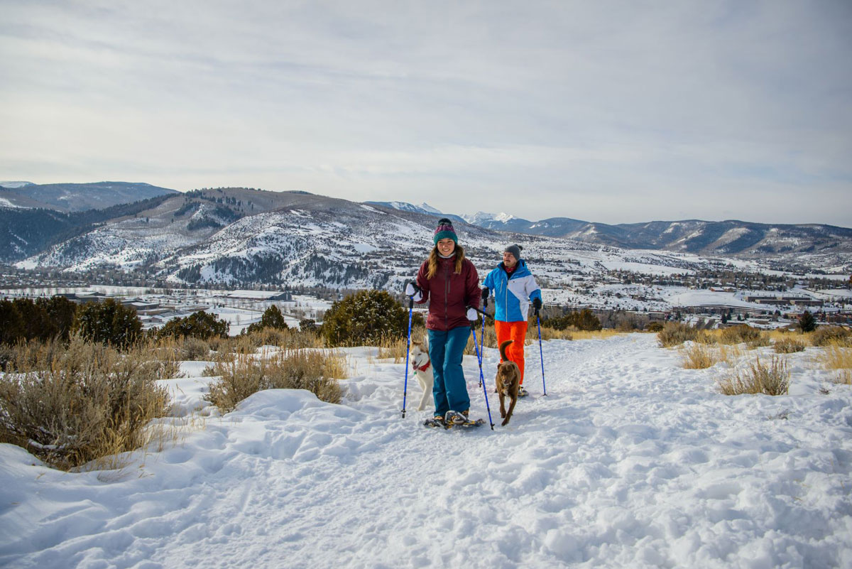Two smiling snowshoers in bright winter clothing walk through the snow-covered hills. Two dogs run beside them happily. In the background are hilly mountains speckled with trees and snow.