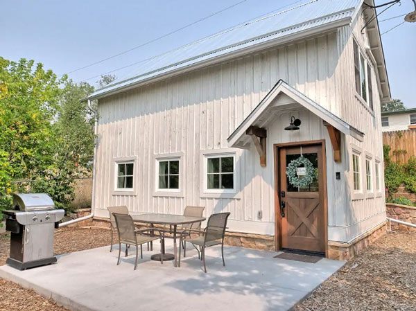 A broad, multi-windowed and tall white barn with a wreath-adorned brown door sits among a patio and some small wood chippings. Around the barn, which is the Dyer Inn at Castle Rock, are various trees and shrubbery.