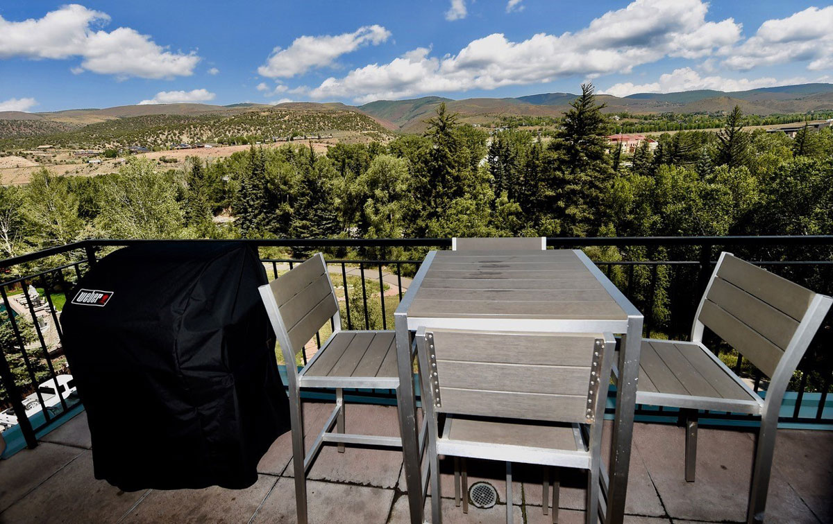 A photo of the balcony at the two bedroom river view condo at The Inn at Riverwalk. There's a covered grill next to the four-chaired table. The view in the distance is that of pine trees and distant rolling mountains. The sky is partly cloudy but the sun is shining.