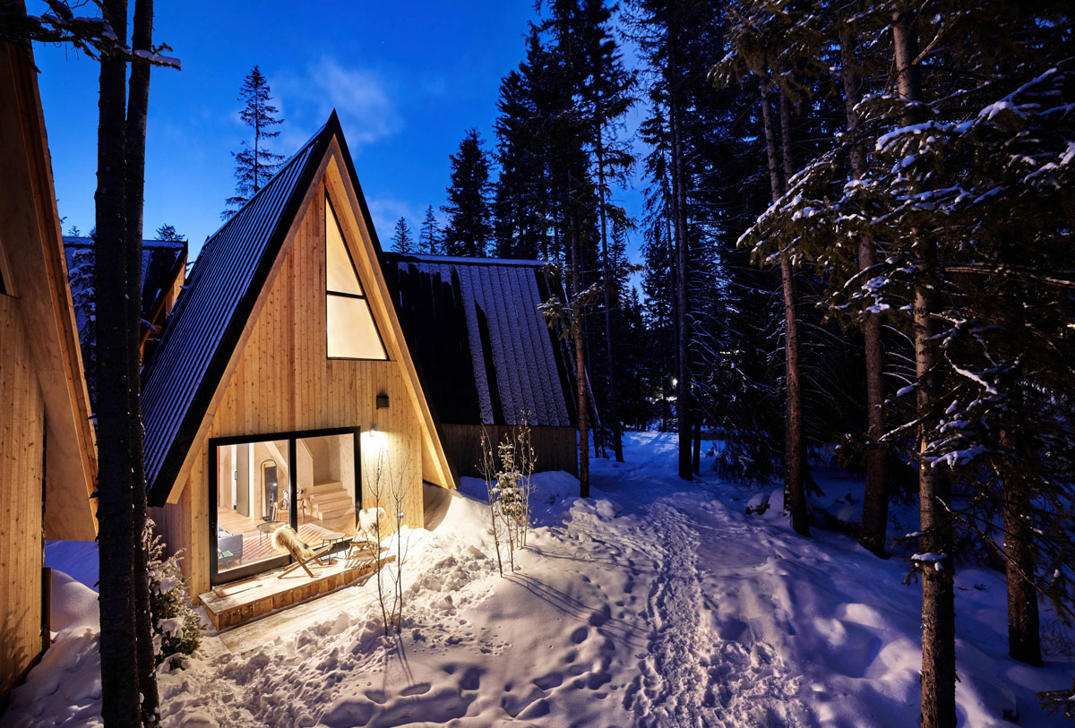 An A-Frame Club cabin is lit up and seen from the exterior surrounded by snow and trees. The night sky is indigo.