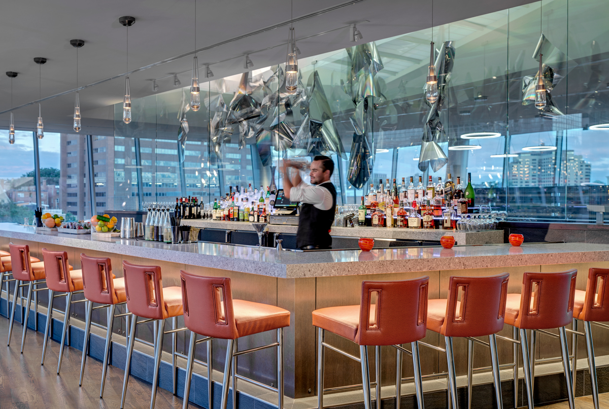A bartender behind The Art's Fire Bar shakes a cocktail in a silver shaker. Views of Denver at sunset are seen from the bar's windows.