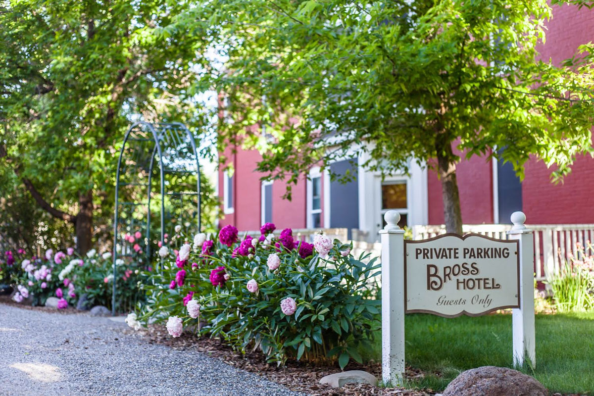 The crimson Bross Hotel Bed & Breakfast is in the background, with a sign, verdant tree leaves and blooming peonies in shades of pink.