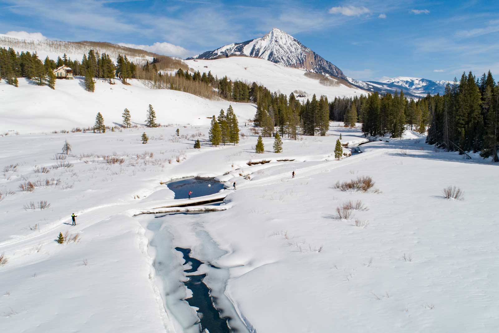 Three cross-country skiers cross a half-frozen river on a bridge in a wide-open snowy meadow