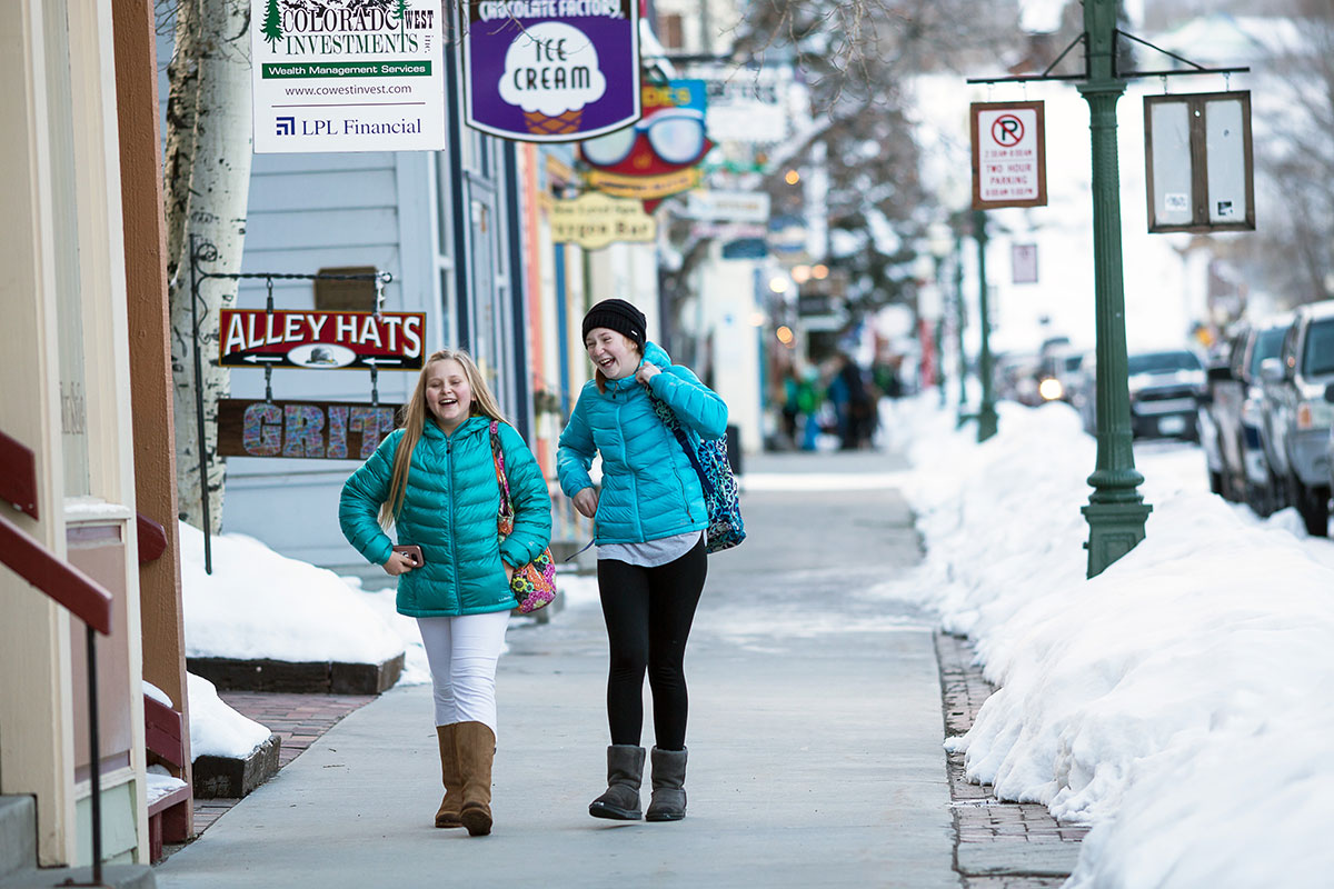 Two young girls laugh and walk down a street with signs for businesses overhead