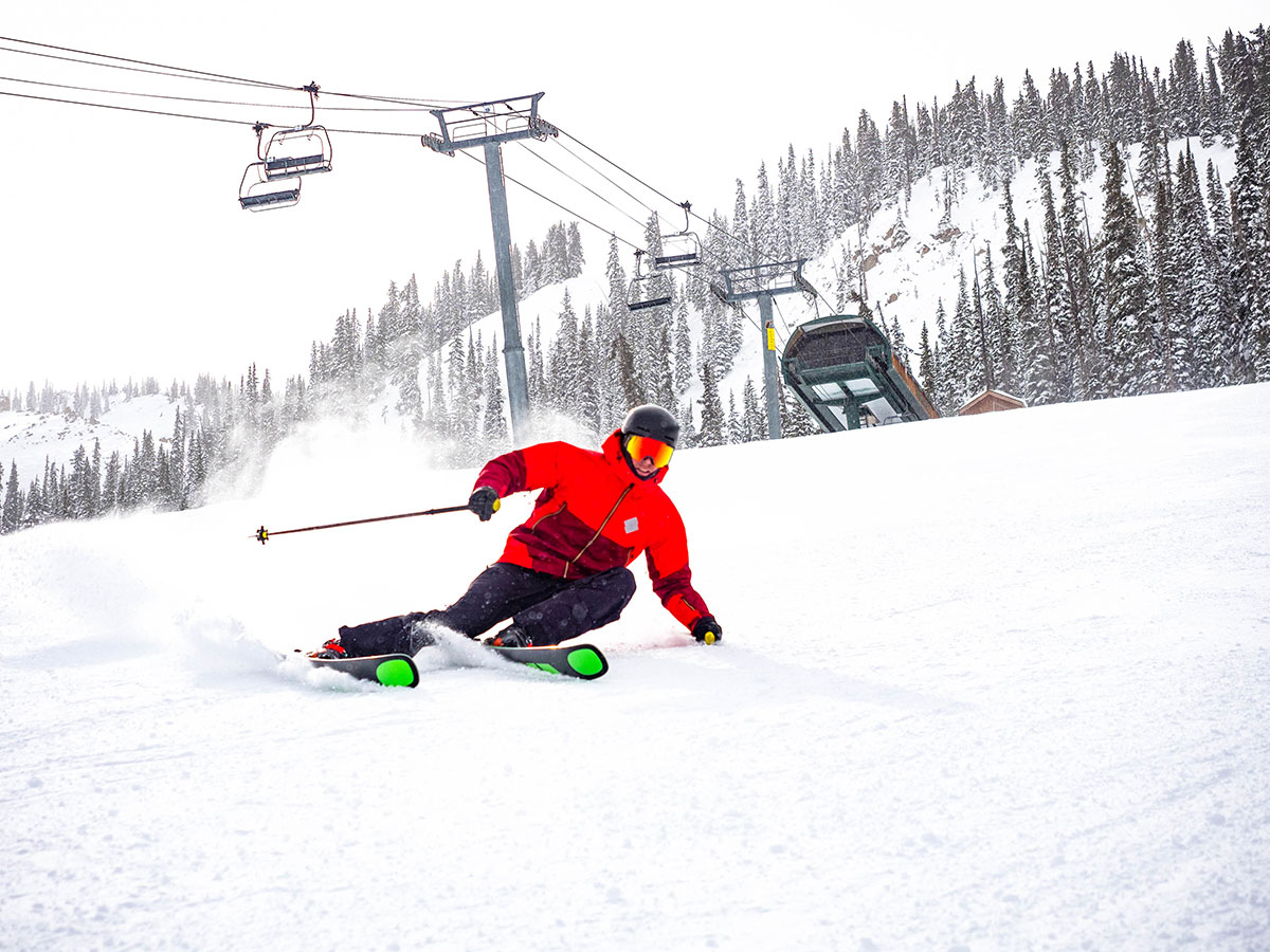 Male skier in a bright-red jacket skiing underneath a ski lift on a mountain