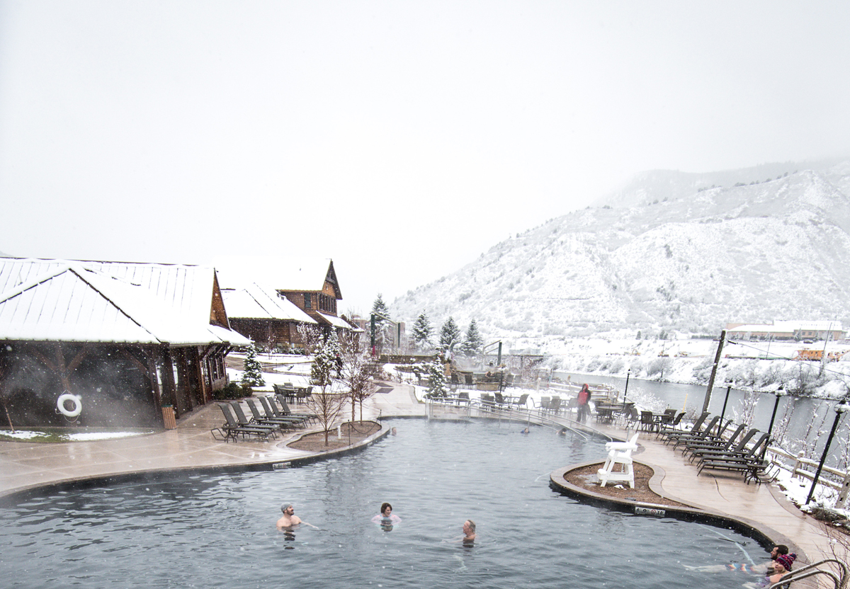 People relax in a large, steaming hot springs pool with a backdrop of snow-blanketed mountains at Iron Mountain Hot Springs in Glenwood Springs, Colorado