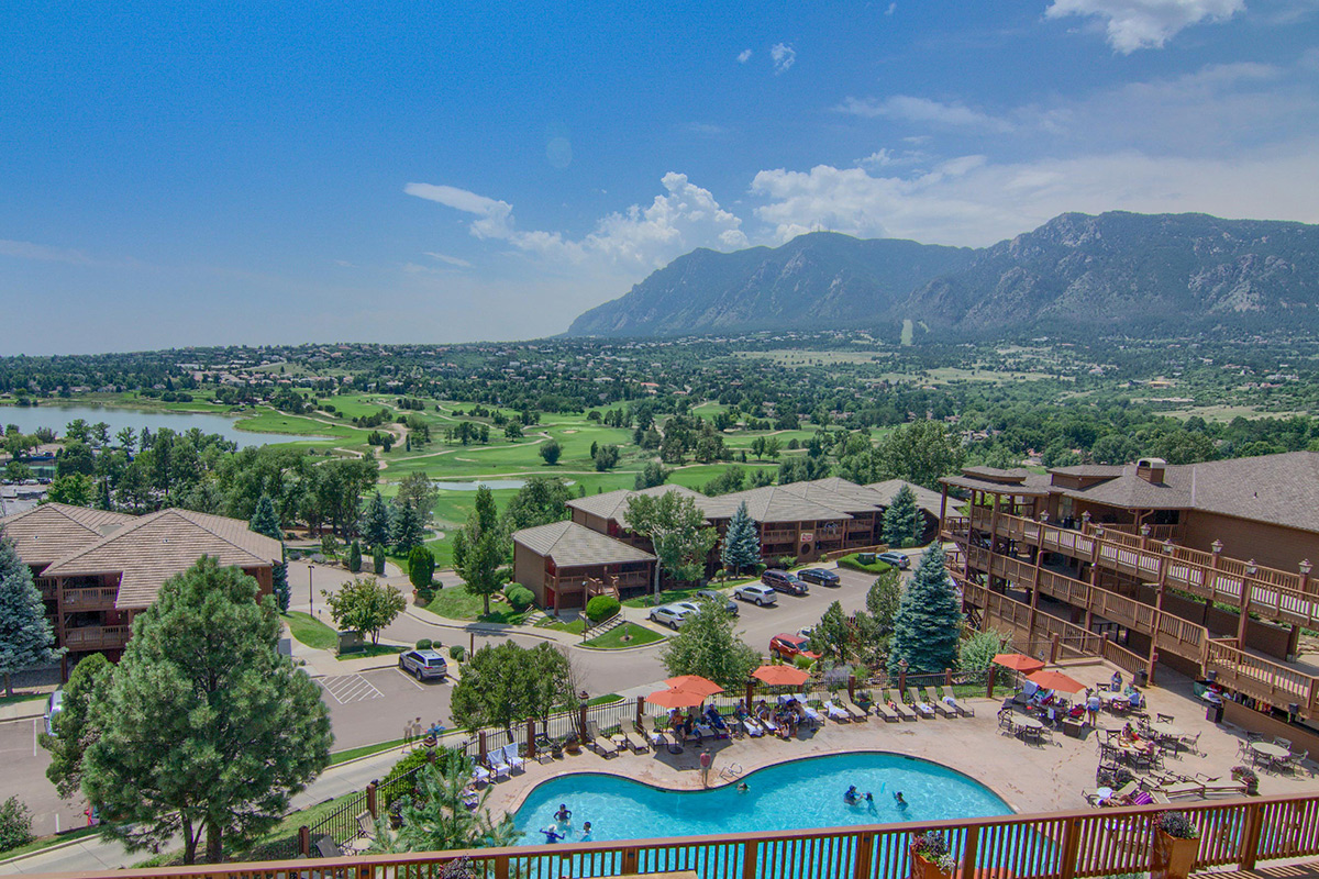 The view from a balcony at Cheyenne Mountain Resort showcases a crystal-clear swimming pool, along with a blue lake, lush golf course and towering peaks in the distance.