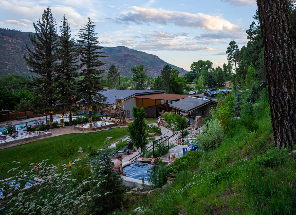people of all ages gather in various hot-springs pools surrounded by blooming summer flowers and mountain views at Durango Hot Springs in Colorado