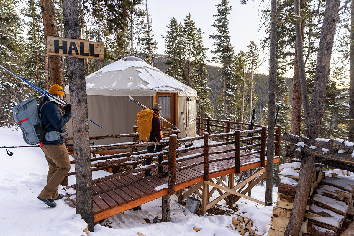 Two people dresses in winter clothing walk through snow onto the deck of a yurt at Tennessee Pass Sleep Yurts in Leadville, Colorado.
