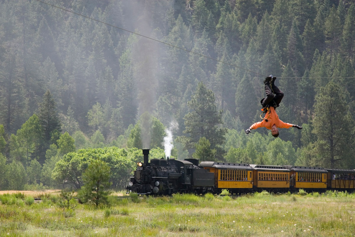 A sky ranger performs an upside-down trick on a zipline with a train in the background in Colorado.