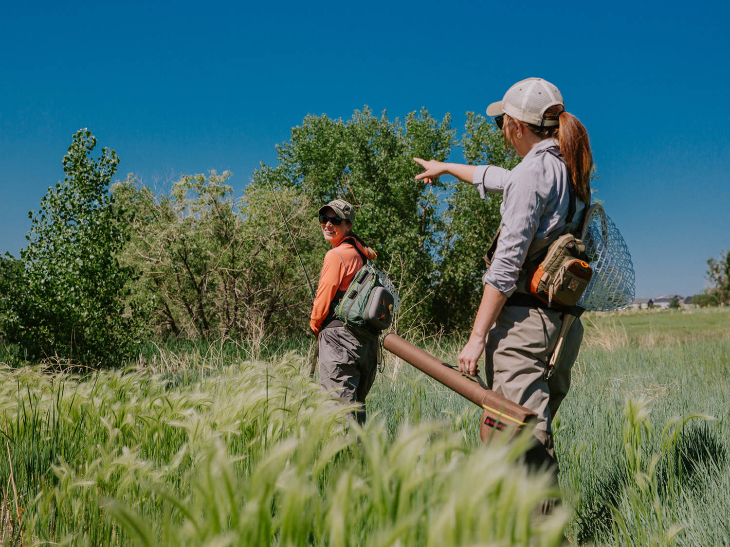 Two people hike through knee-high grass carrying fly-fishing gear. 