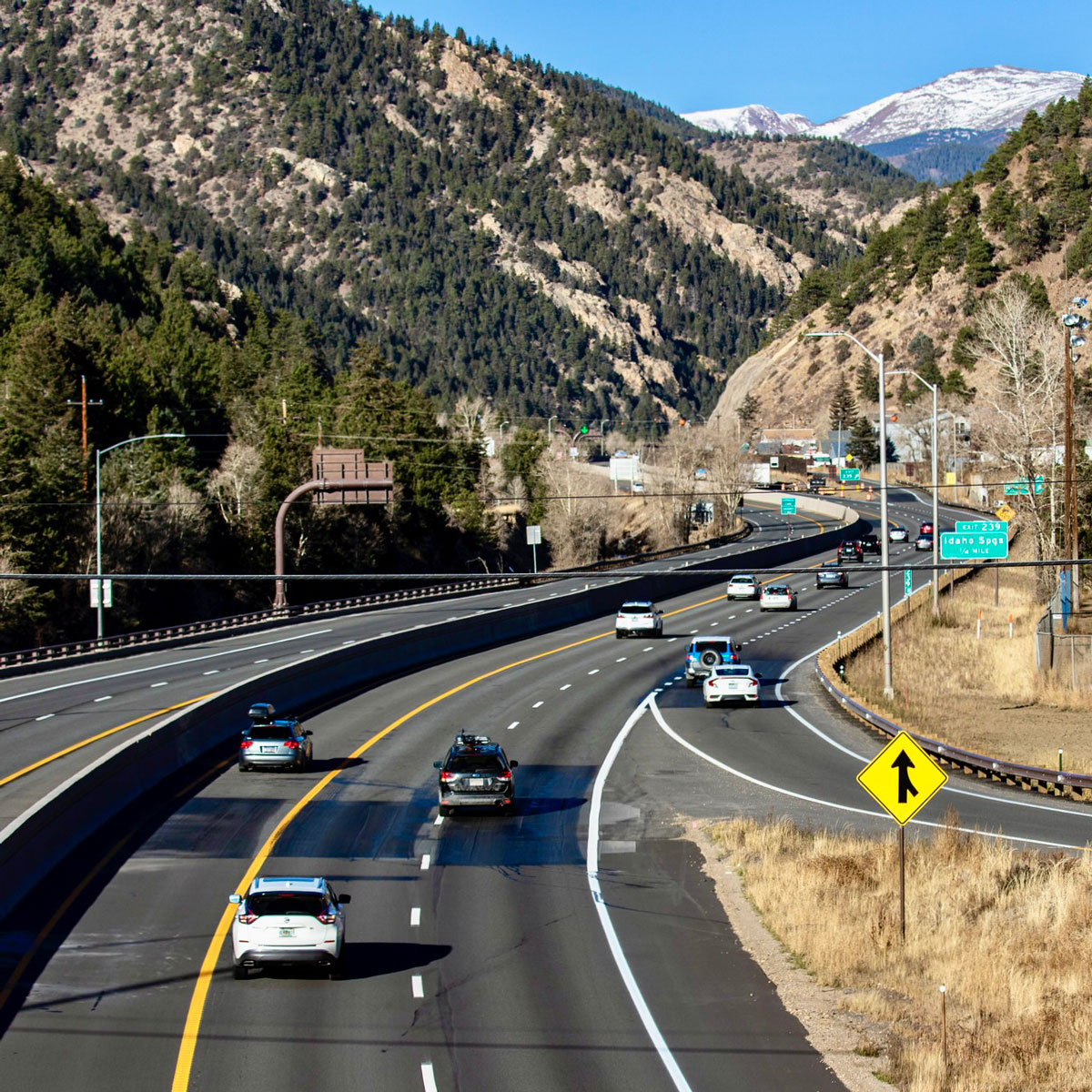 Vehicles driving along the highway, surrounded by towering forested and snow-covered mountains in Colorado.