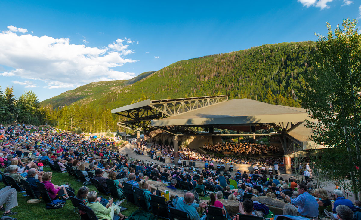 Concert goers sit on the lawn in front of a stage backed by green forest