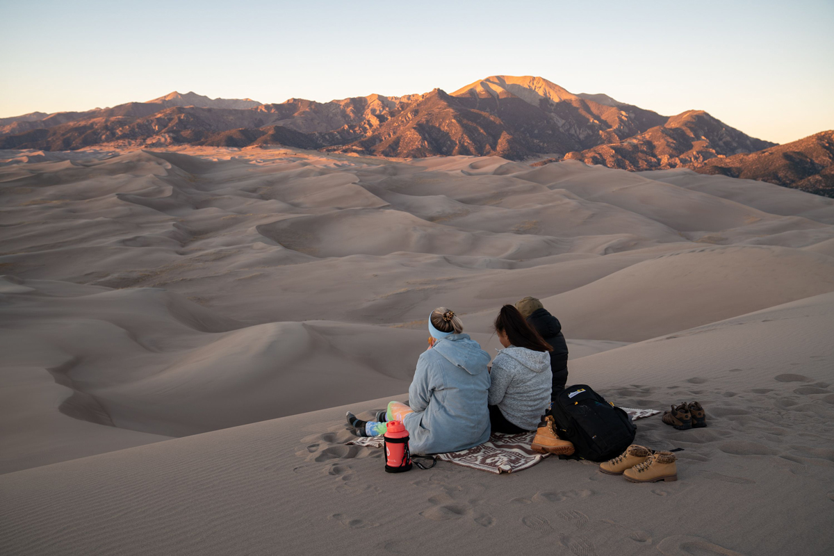 Two people sit closely on the dunes of Great Sand Dunes National Park and Preserve and look at the bordering Sangre de Cristo Mountains in Alamosa Colorado