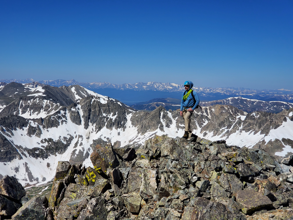 A person stands on a snow-streaked ridge in mountaineering gear with peaks rising in the distance below in the background on a guided mountaineering trip with Colorado Adventure Guides. 