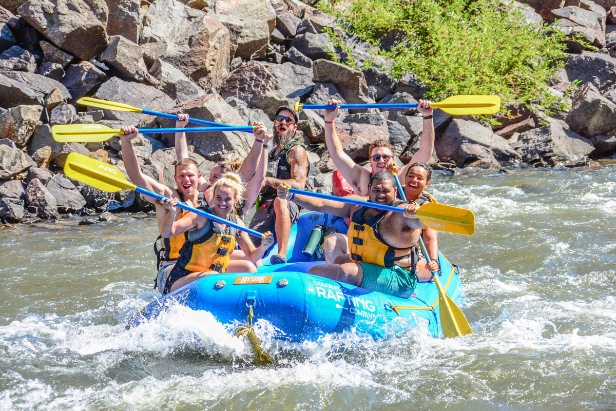 A blue raft filled with people wearing life jackets and helmets raise their paddles and cheer while passing through a section of whitewater.