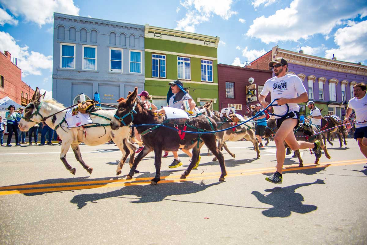 people and donkeys run down a Leadville street with colorful buildings along the sides