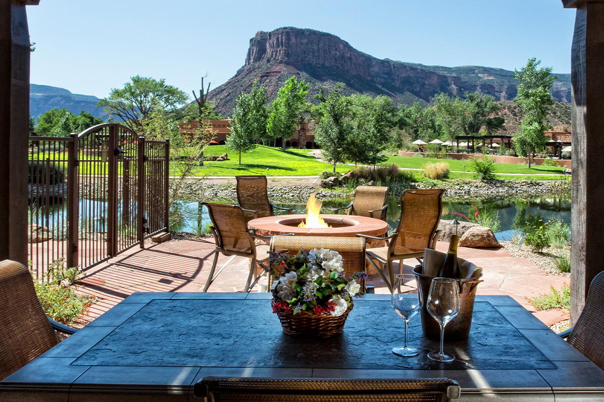 At Gateway Canyon Resort in Gateway, Colorado, a firepit surrounded by chairs and a table set with wine are framed by a tranquil pond and a canyon wall rising in the distance.