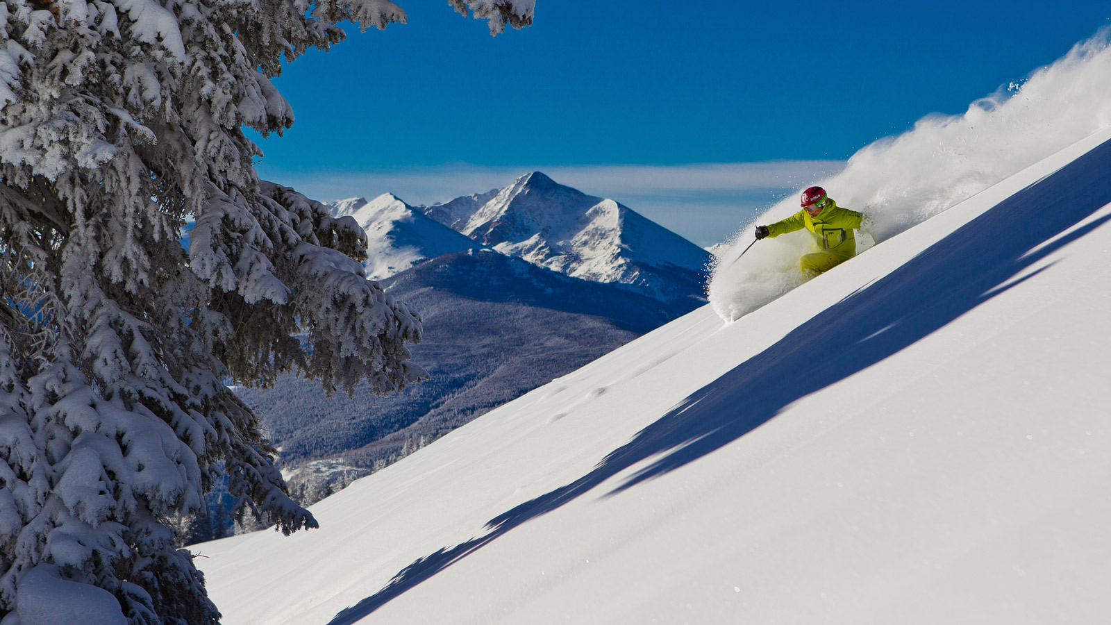 A skier blows up some powder as they charge downhill under sunny Colorado skies