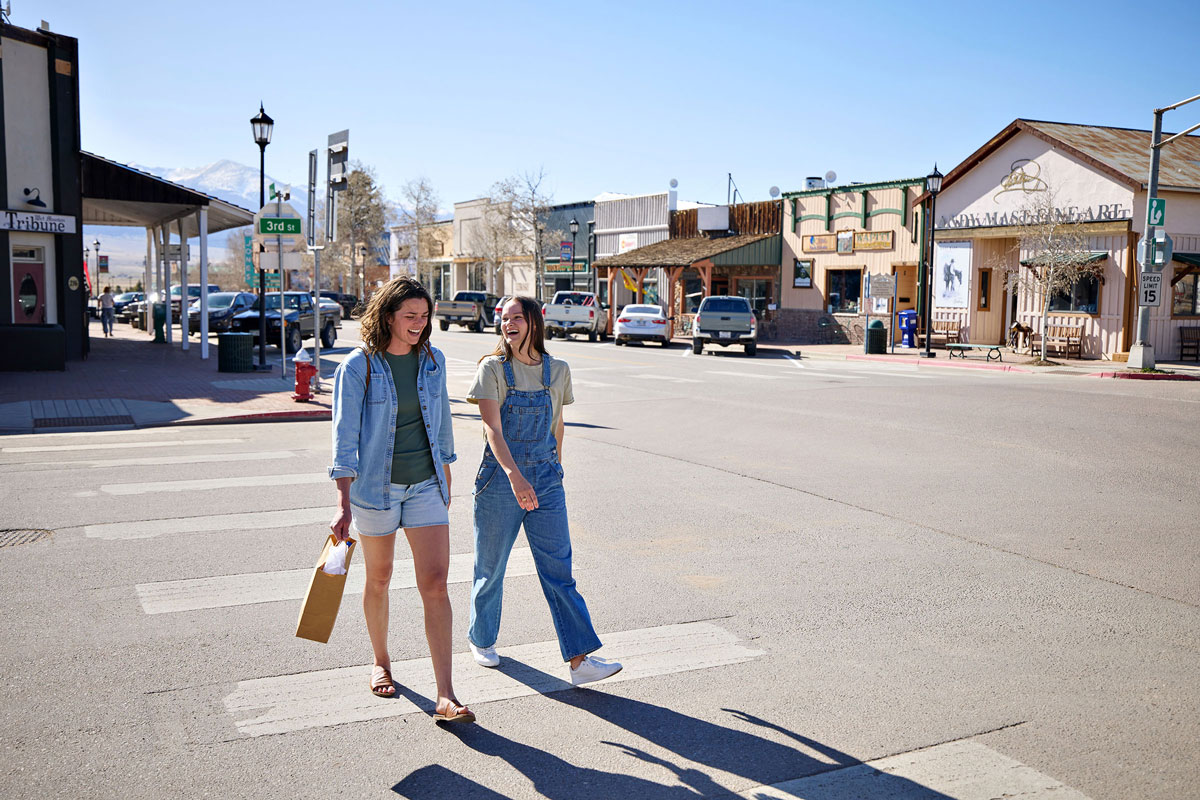 Two girlfriends crossing a road in downtown Westcliffe with a shopping bag in hand and stores behind them.