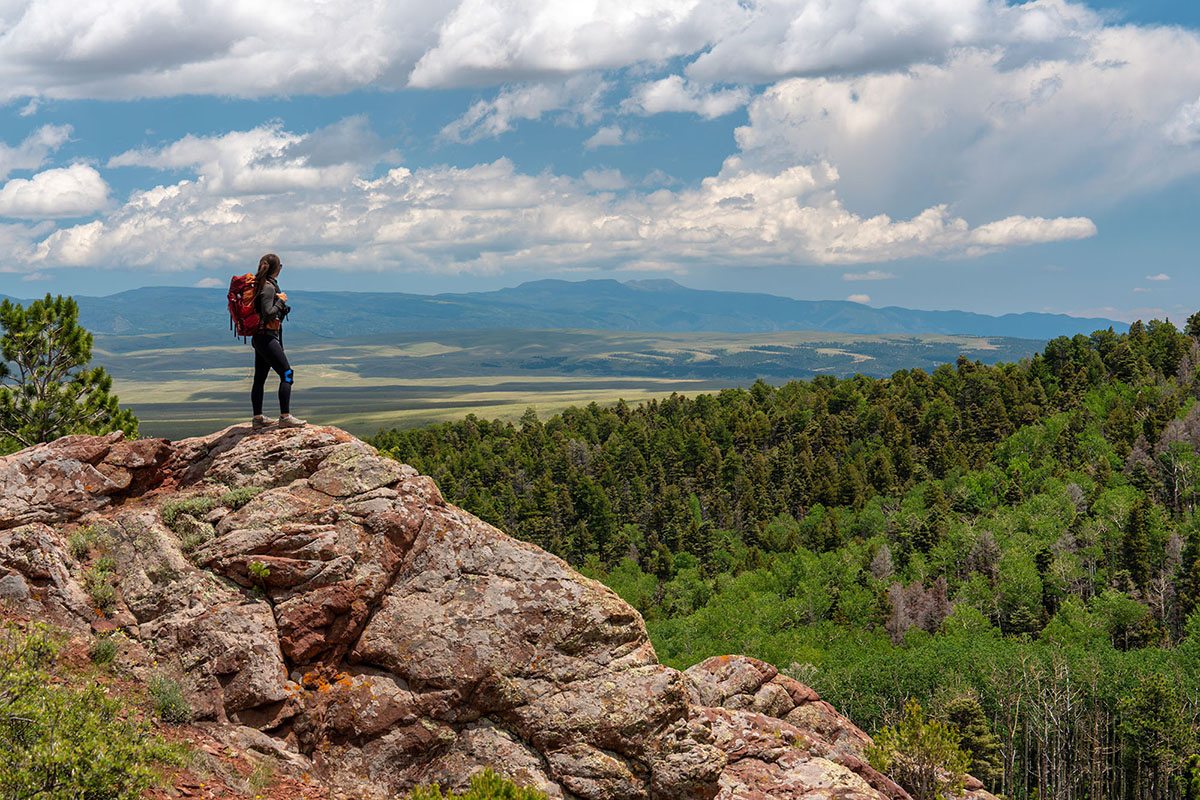 A hiker stands on a rocky outcropping overlooking a forest and valley along the Rainbow Trail near Westcliffe, Colorado