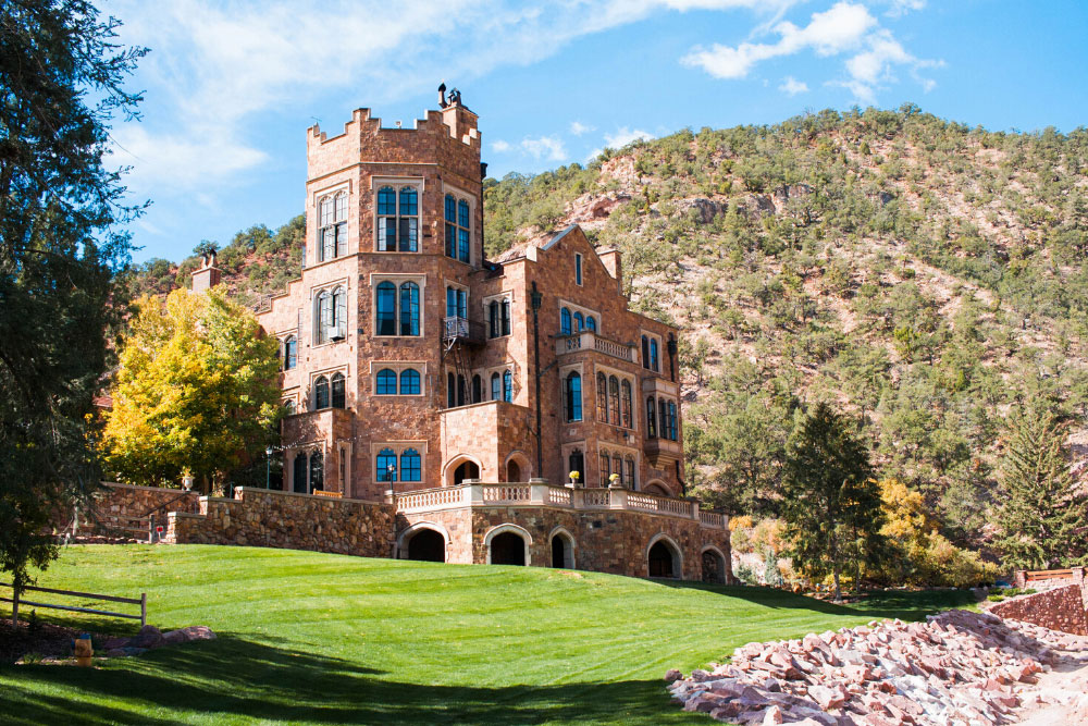 The historical Glen Eyrie Castle in Colorado Springs with stone towers, a green lawn and Rocky Mountain foothills in the background. 